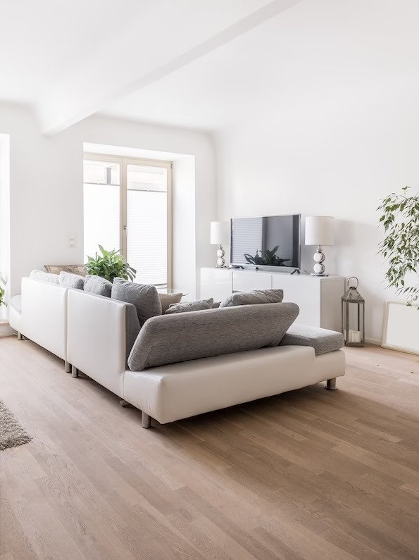Modern living room featuring a white sectional sofa, light hardwood flooring, a television on a sleek console, and natural light from large windows, showcasing a clean and contemporary aesthetic ideal for flooring installation services.