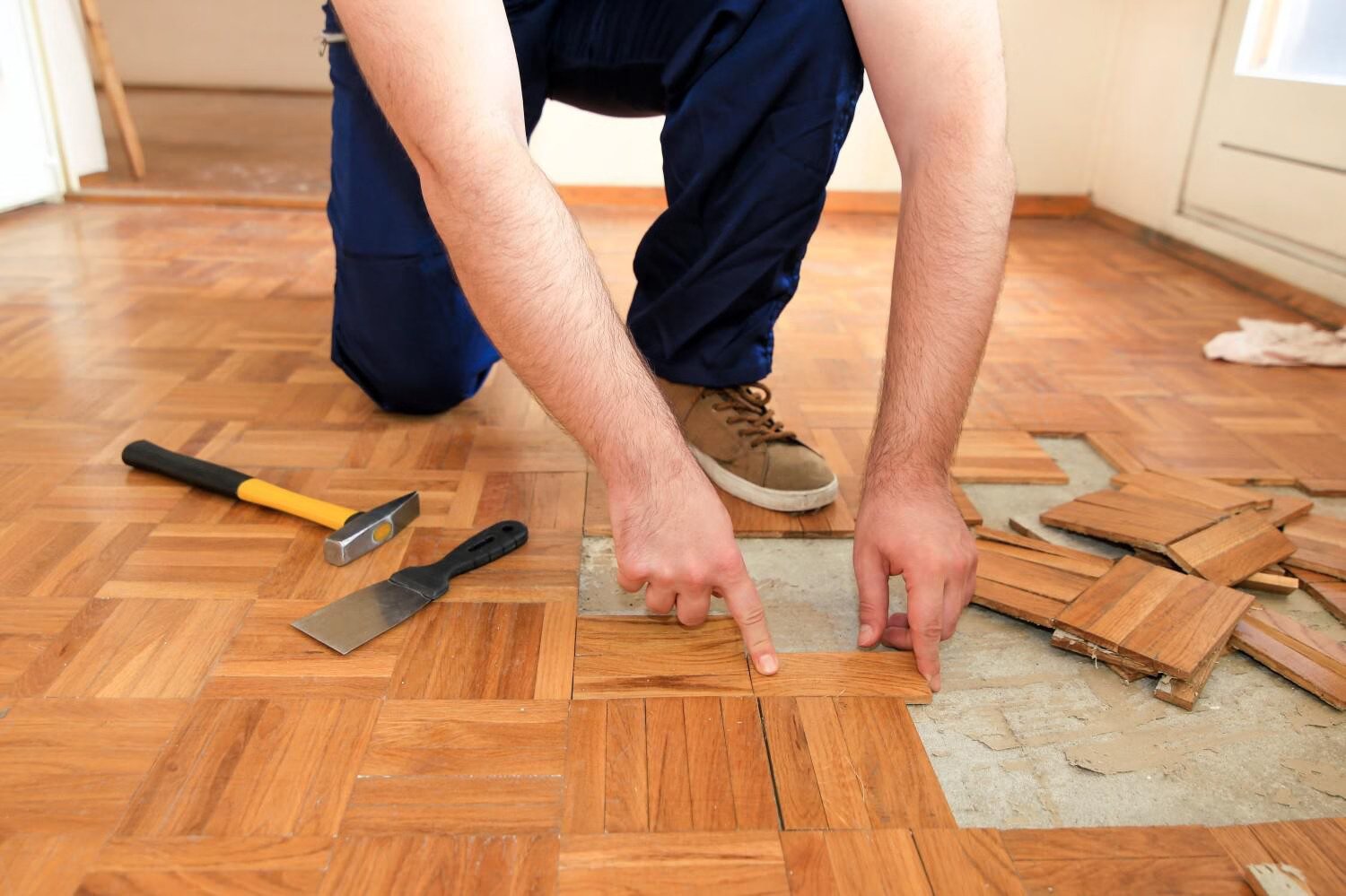 Person kneeling on hardwood floor repairing parquet tiles, pointing at a section while holding tools, demonstrating floor repair services.