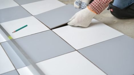 Person installing tile flooring with gray and white tiles, using a pencil for measurement, emphasizing professional tile installation services for kitchens and bathrooms.